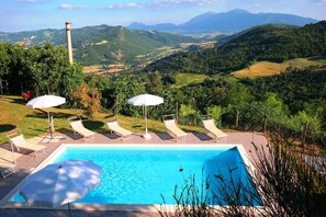 Ein Schwimmbecken mit Liegestühlen und Sonnenschirmen, umgeben von üppiger Landschaft und Bergen im Hintergrund.