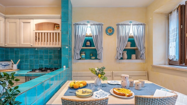 Een keuken met een blauwe betegelde achterwand, witte kasten en een eettafel gedekt met borden, glazen en een vaas met bloemen.