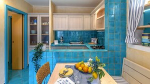 Een keuken met blauwe tegels, een eettafel met een fruitschaal, en een rieten stoel.