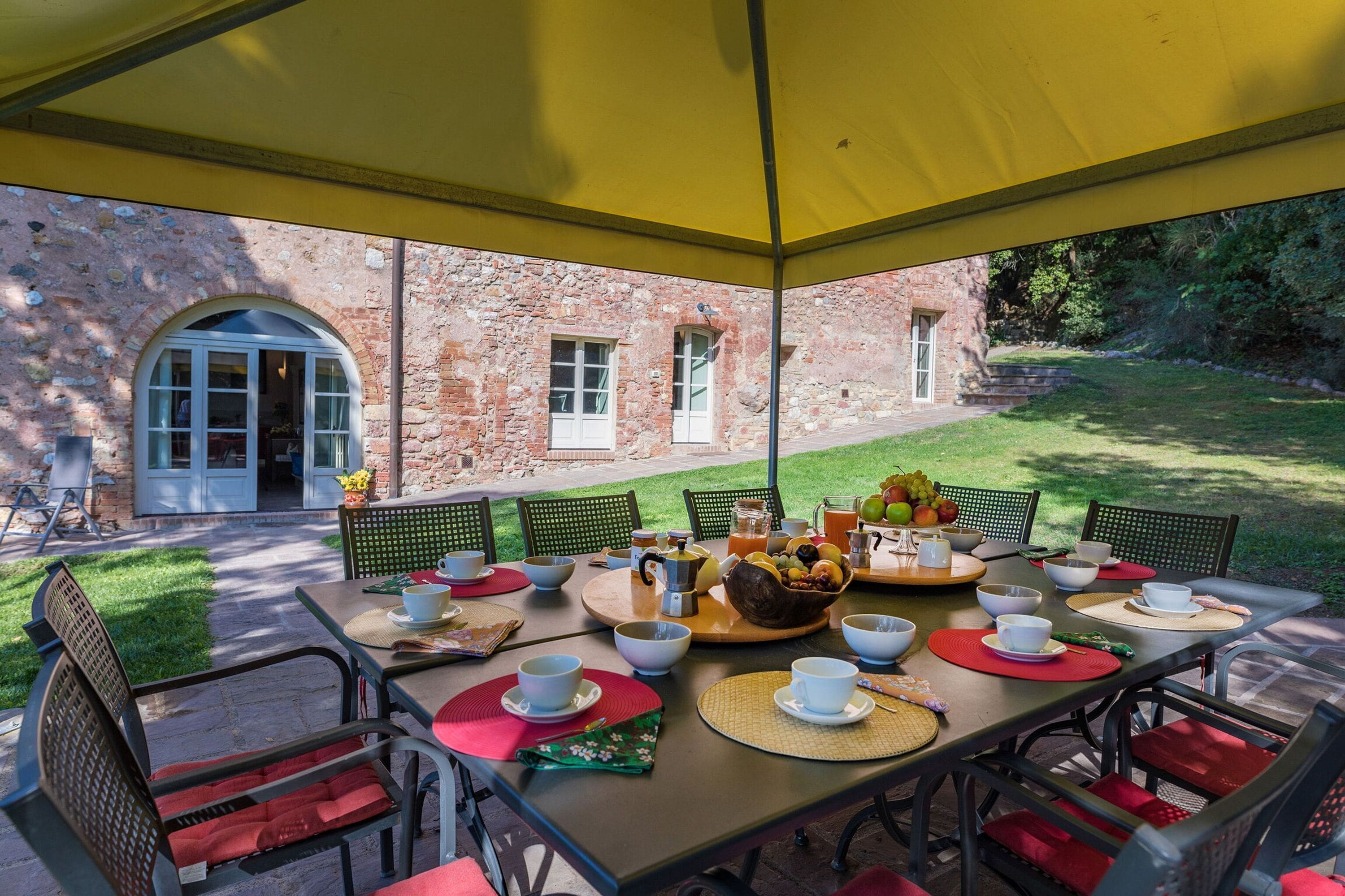 An outdoor dining area with a table set for breakfast, surrounded by chairs and a yellow canopy.