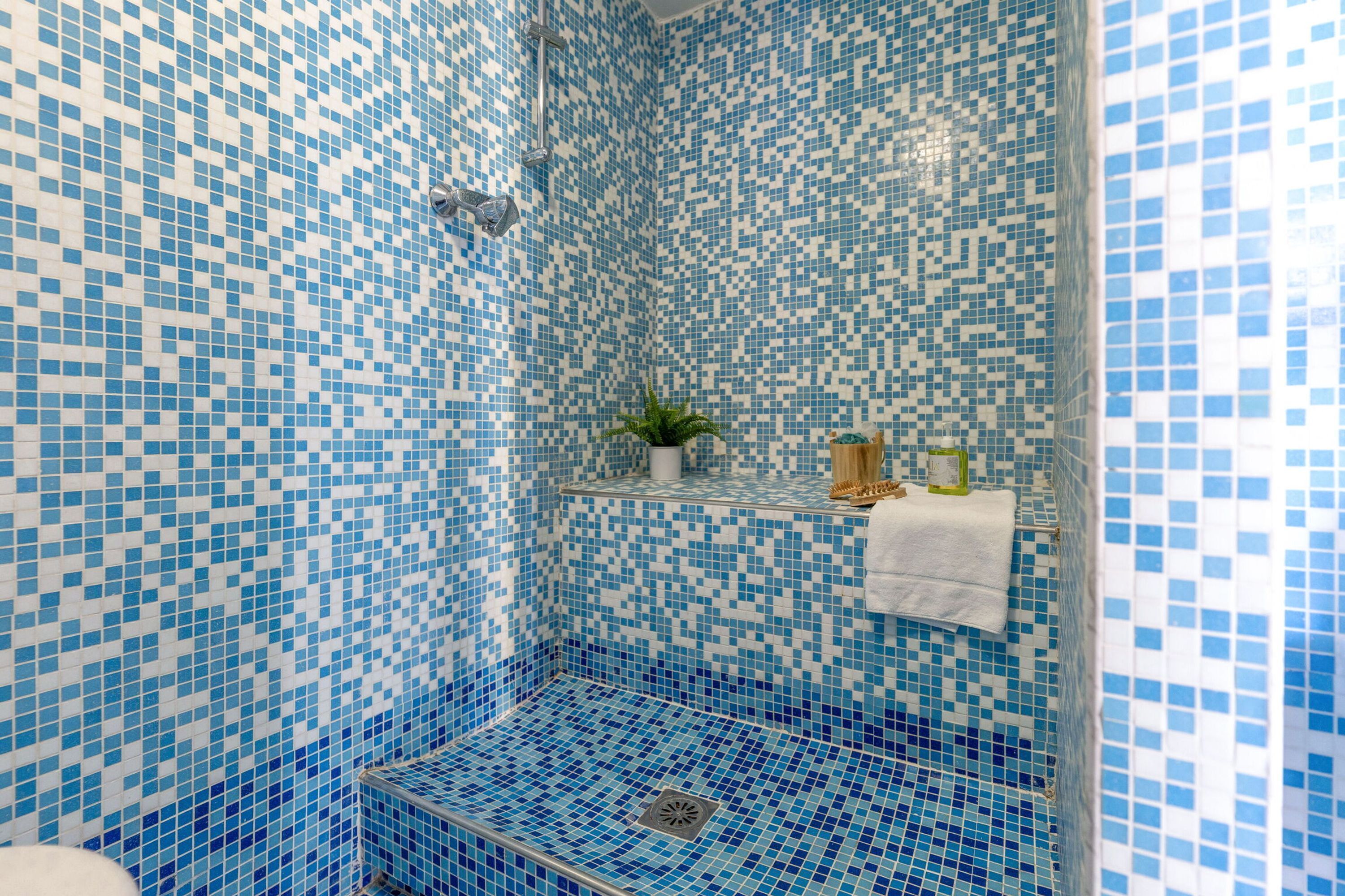 A bathroom with blue mosaic tiles, a shower, a sink with a plant, and a towel rack.