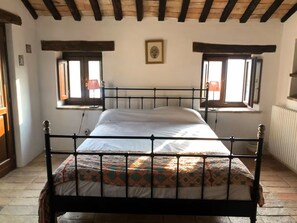 Una habitación con cama, dos ventanas, radiador y vigas de madera en el techo.