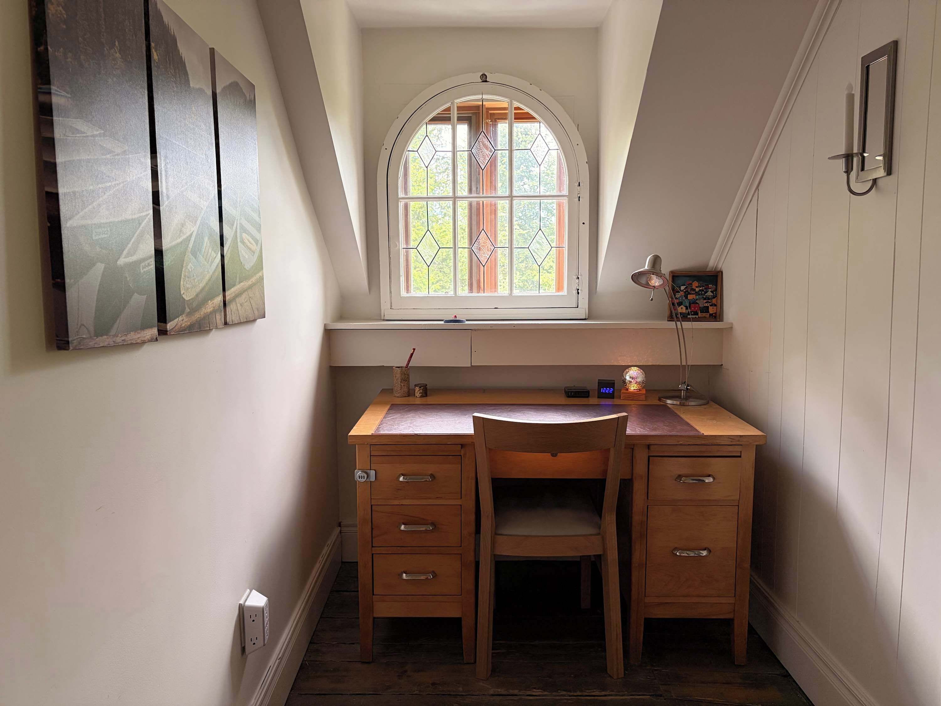 A small, cozy study room with a wooden desk, two chairs, a window with stained glass, and a wall-mounted lamp.