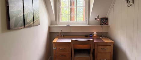A small, cozy study room with a wooden desk, two chairs, a window with stained glass, and a wall-mounted lamp.