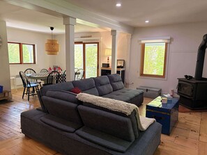 A modern living room with a grey sectional sofa, a dining area with a table and chairs, a balcony with a view, and a wood-burning stove. - L'Ancestrale de Fitch Bay (Magog)