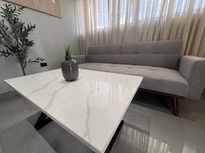 Una sala de estar moderna con una mesa de centro de mármol blanco, un jarrón gris con una planta, un sofá gris y un ventanal con cortinas traslúcidas.