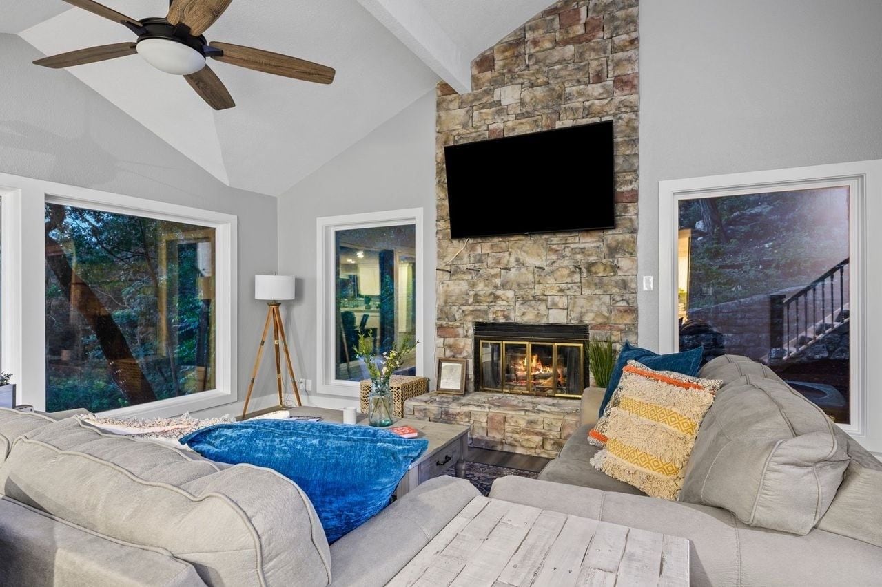 A living room with a stone fireplace, a ceiling fan, and a large window.