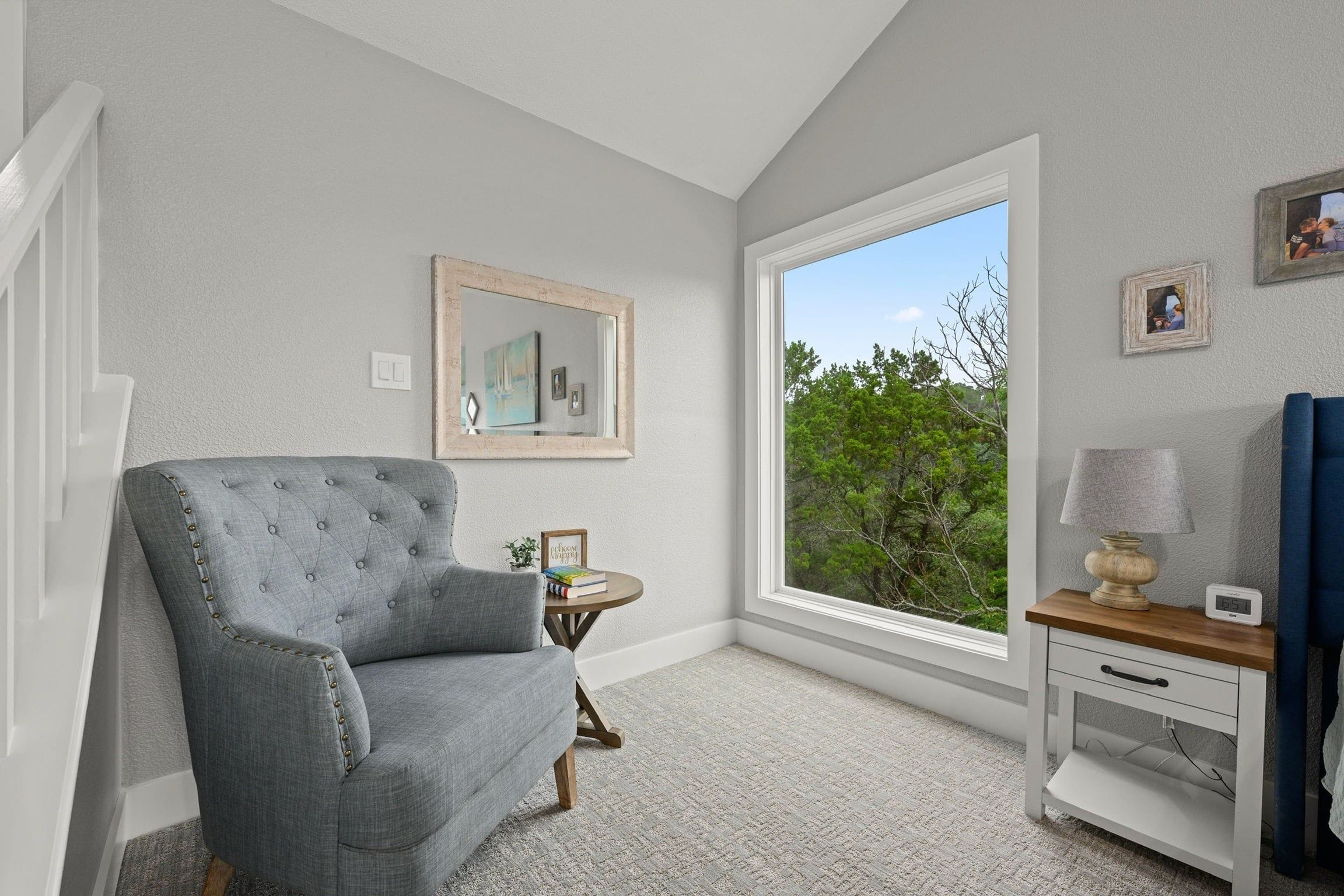 A room with a grey upholstered armchair, a small wooden side table, a white dresser, a large window with a view of trees, and a framed mirror on the wall.