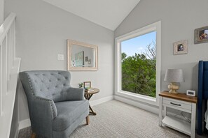 Ein Zimmer mit einem grau gepolsterten Sessel, einem kleinen Holznachttisch, einem weißen Schminktisch und einem großen Fenster mit Blick auf Bäume. An der Wand hängt ein gerahmter Spiegel.