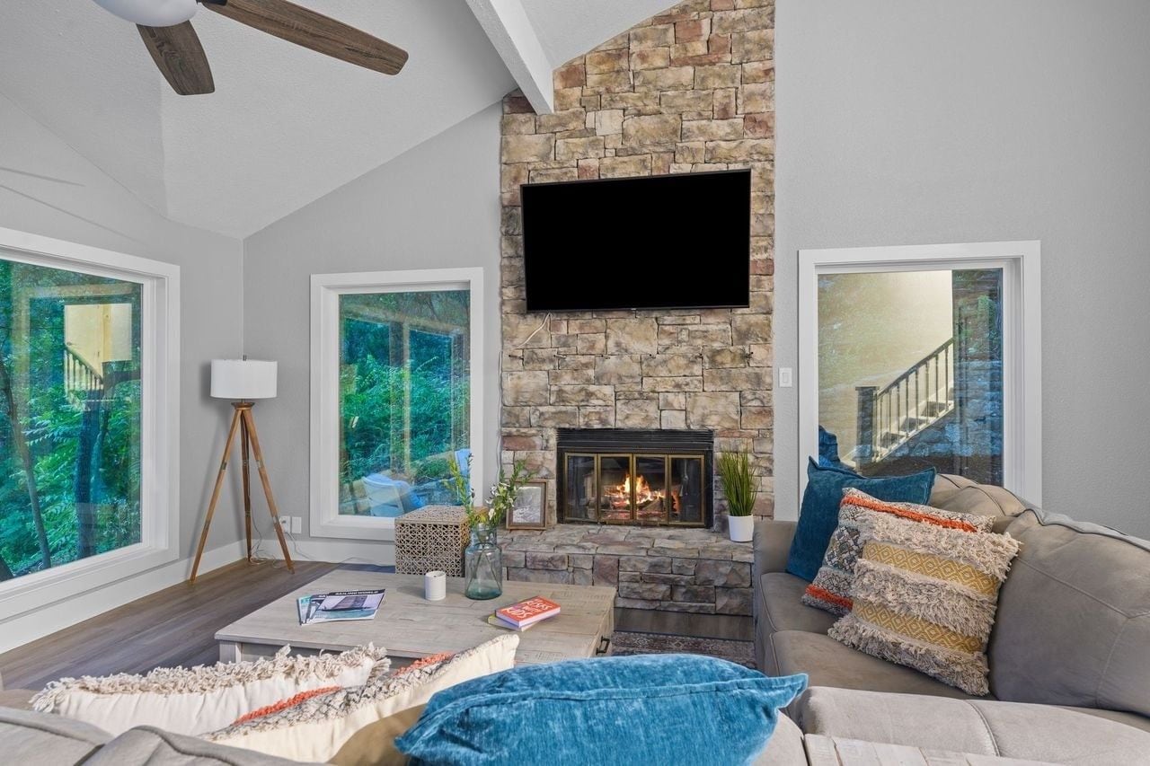 Una sala de estar con chimenea de piedra, ventanas amplias, televisor de pantalla plana y un rincón de estar acogedor.