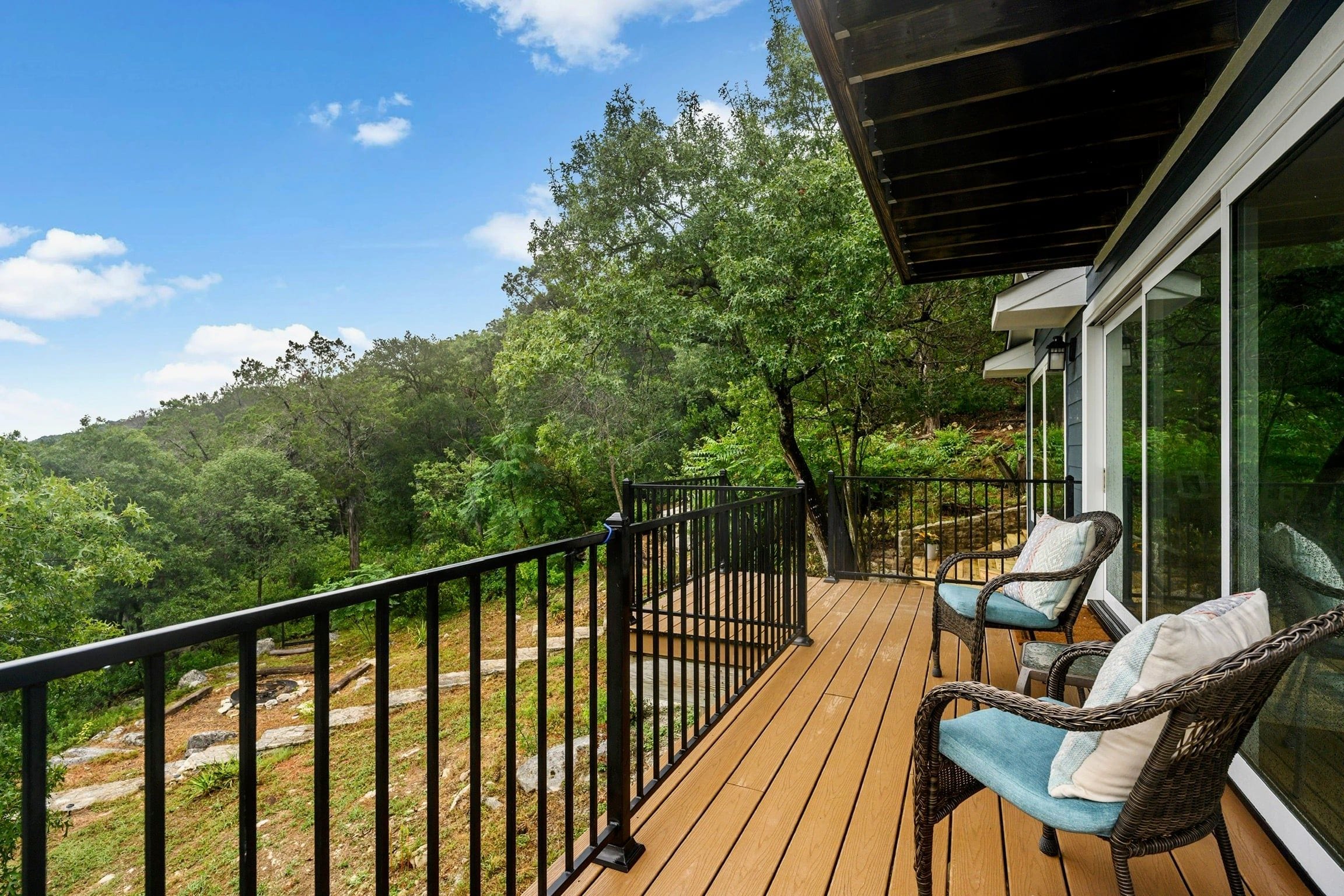 A wooden deck with a black railing, two wicker chairs with cushions, and a glass door leading to a lush green landscape.