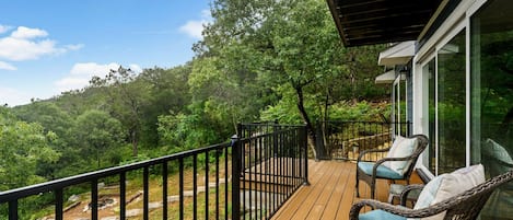 A wooden deck with a black railing, two wicker chairs with cushions, and a glass door leading to a lush green landscape.