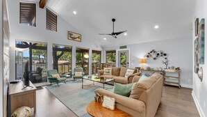 Uma sala de estar ampla com um sofá grande, uma mesa de centro e uma televisão. Há várias janelas que oferecem luz natural e vista para o exterior.