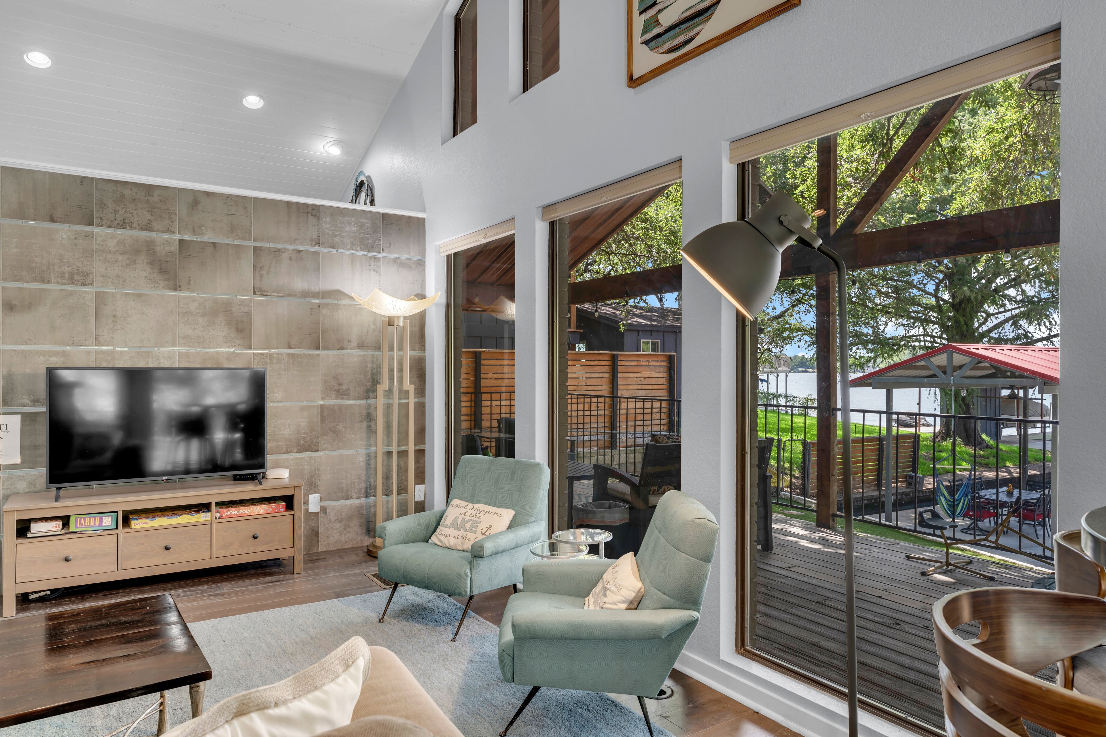 A modern living room with a flat-screen TV, comfortable seating, and large windows offering a view of the outdoors.