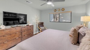 Quarto com cama, cômoda, televisão, ventilador de teto, janela e decorações nas paredes.