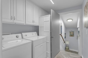 A laundry area with two front-loading washers and dryers, a door leading to another room, and a hallway with a lamp and wall art.