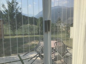 A view through a mesh screen with a patio area, including chairs and tables, and a mountainous landscape in the background.
