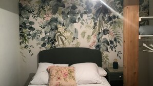 Un letto con biancheria da letto bianca e un cuscino floreale, un comodino con una lampada e una parete con carta da parati a motivi floreali.