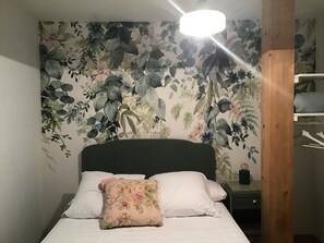 A bed with white bedding and a floral pillow, a bedside table with a lamp, and a wall with a floral wallpaper pattern. - At the foot of the Hautacam climb (Ayros-Arbouix)