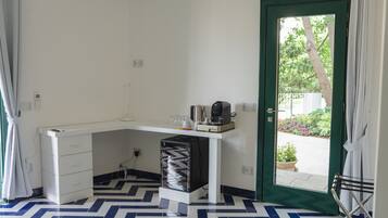 A compact room with a desk, a chair, and a coffee machine. There is a door leading to an outdoor area with plants.