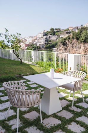 An outdoor patio with a white table and chairs, a white fence, and a view of a hillside with buildings. - Casacasa Positano (Campania)