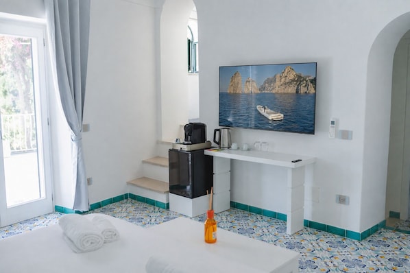 A hotel room with a bed, a desk with a coffee maker, a painting of a boat on water, and a view of the sea through an arched window. - Casacasa Positano (Campania)