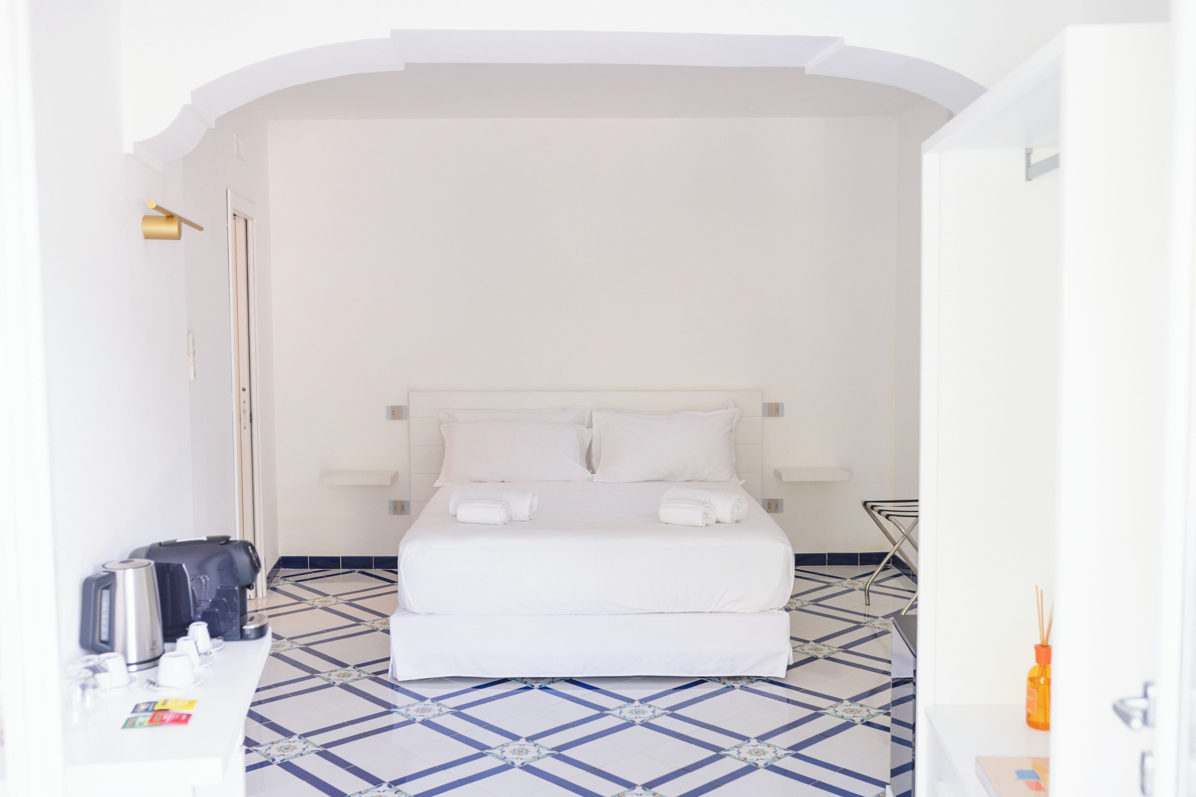 A hotel room with a bed, a bedside table, a chair, a lamp, and a tiled floor.