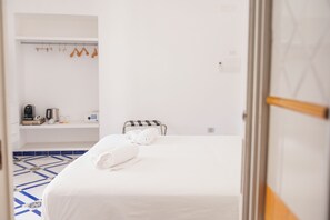 Una camera d'albergo con un letto, un asciugamano, un tavolino con una macchina per il caffè e uno scaffale con oggetti appesi.