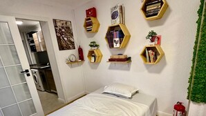 A bedroom with a bed, a small shelf with books, a wall clock, and a fire extinguisher.