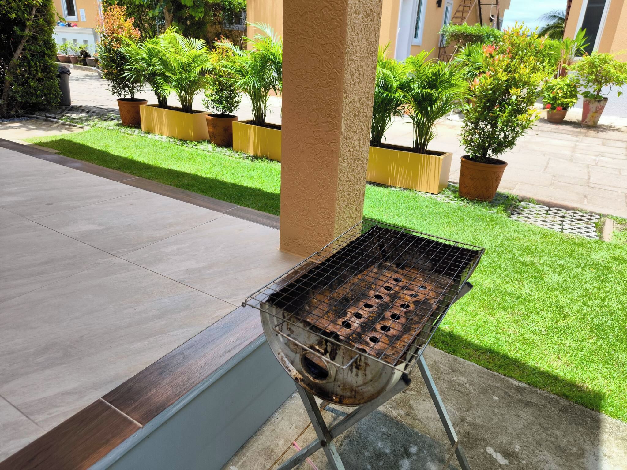 Ein Grillbereich im Freien mit einem Metallrost, einem Betonboden und einem Rasen mit Blumentöpfen.