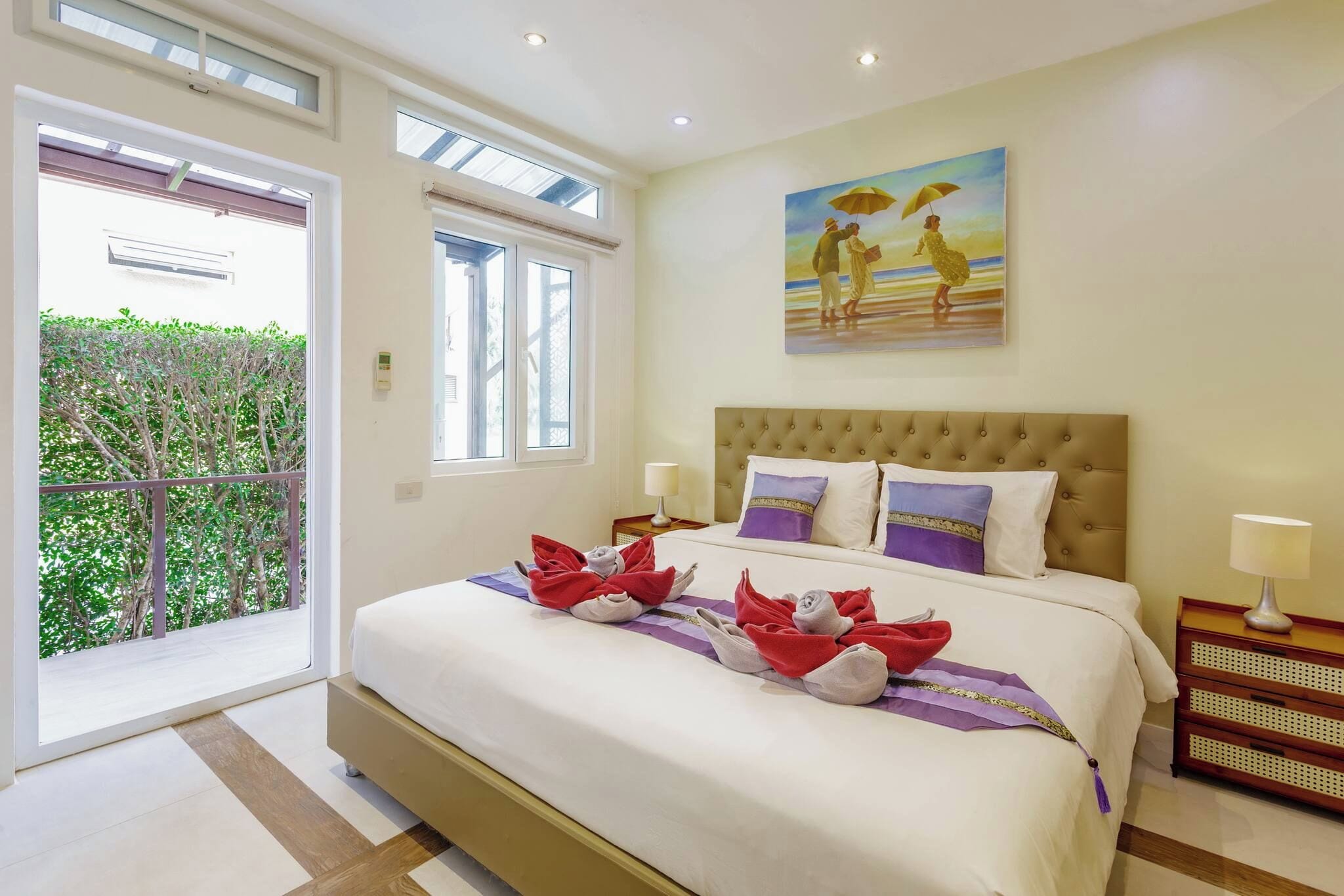 A hotel room with a large bed, a painting on the wall, and a balcony with a view of greenery.