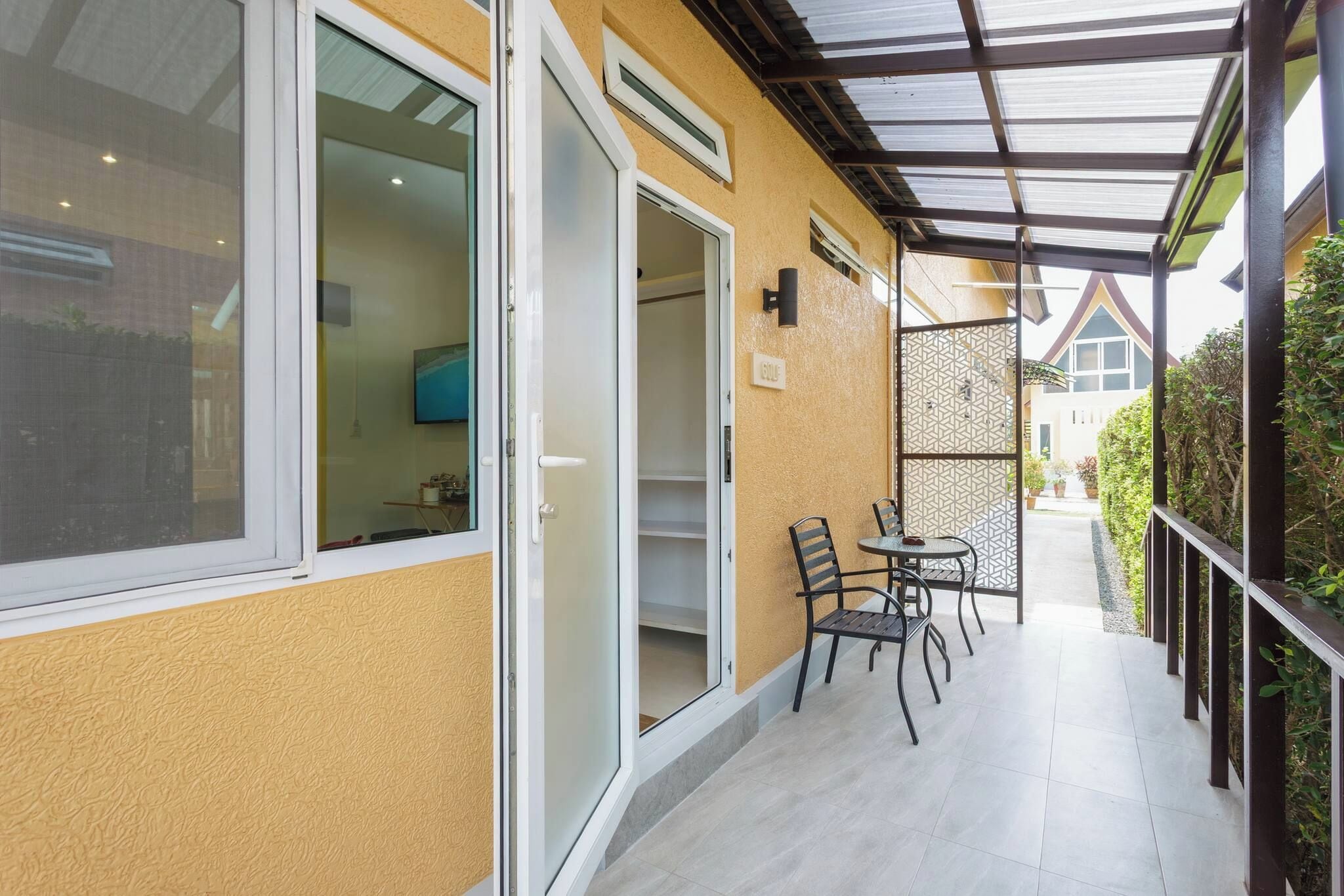 A covered outdoor area with a table and chairs, a door leading to a room, and a view of a building in the background.