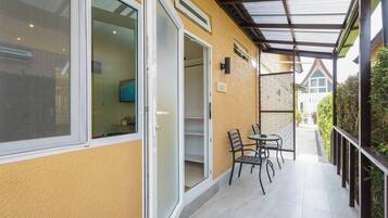 A covered outdoor area with a table and chairs, a door leading to a room, and a view of a building in the background.