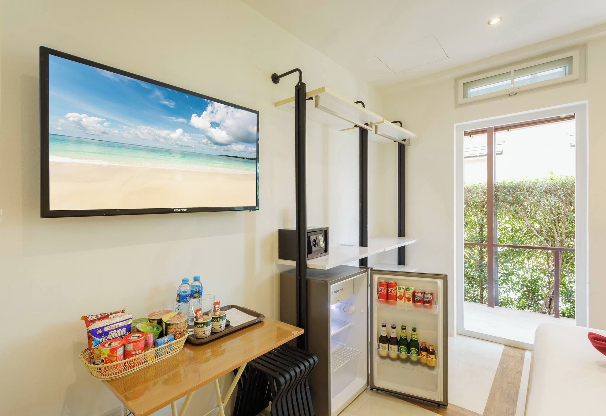 A room with a television displaying a beach scene, a small refrigerator stocked with beverages, a table with snacks and a basket, and a window with a view of greenery.