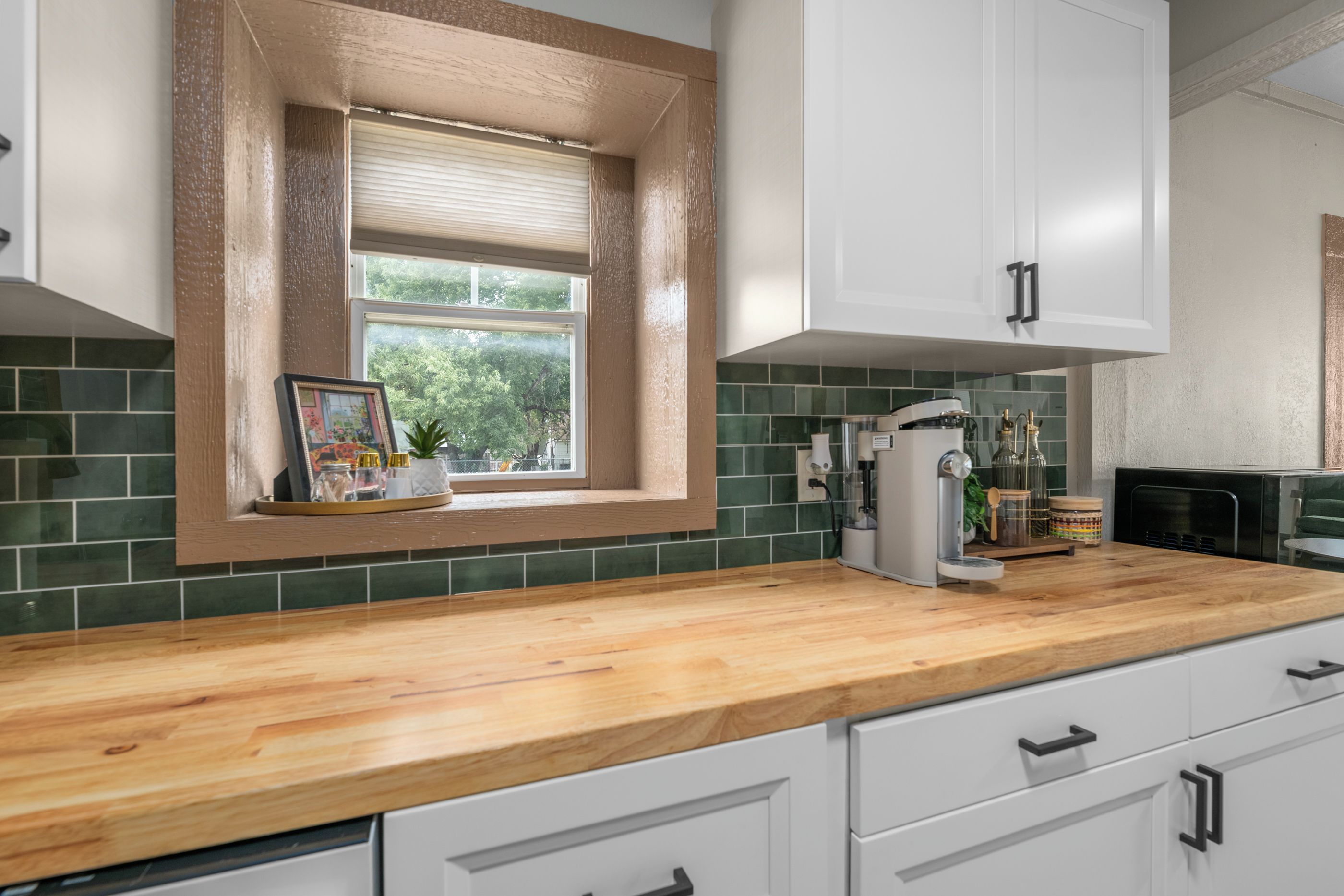 A kitchen with a window, green tiles, a wooden countertop, and white cabinets.