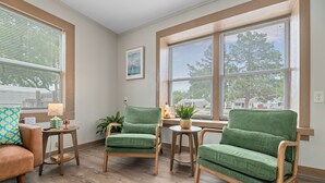 Una habitación con dos sillones verdes, una mesa de madera y una lámpara. Hay una ventana con persianas y un cuadro en la pared.