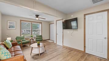 Ein Wohnzimmer mit braunem Ledersofa, grünen Sessel und einem runden Holztisch. Eine Deckenventilator ist ebenfalls vorhanden.