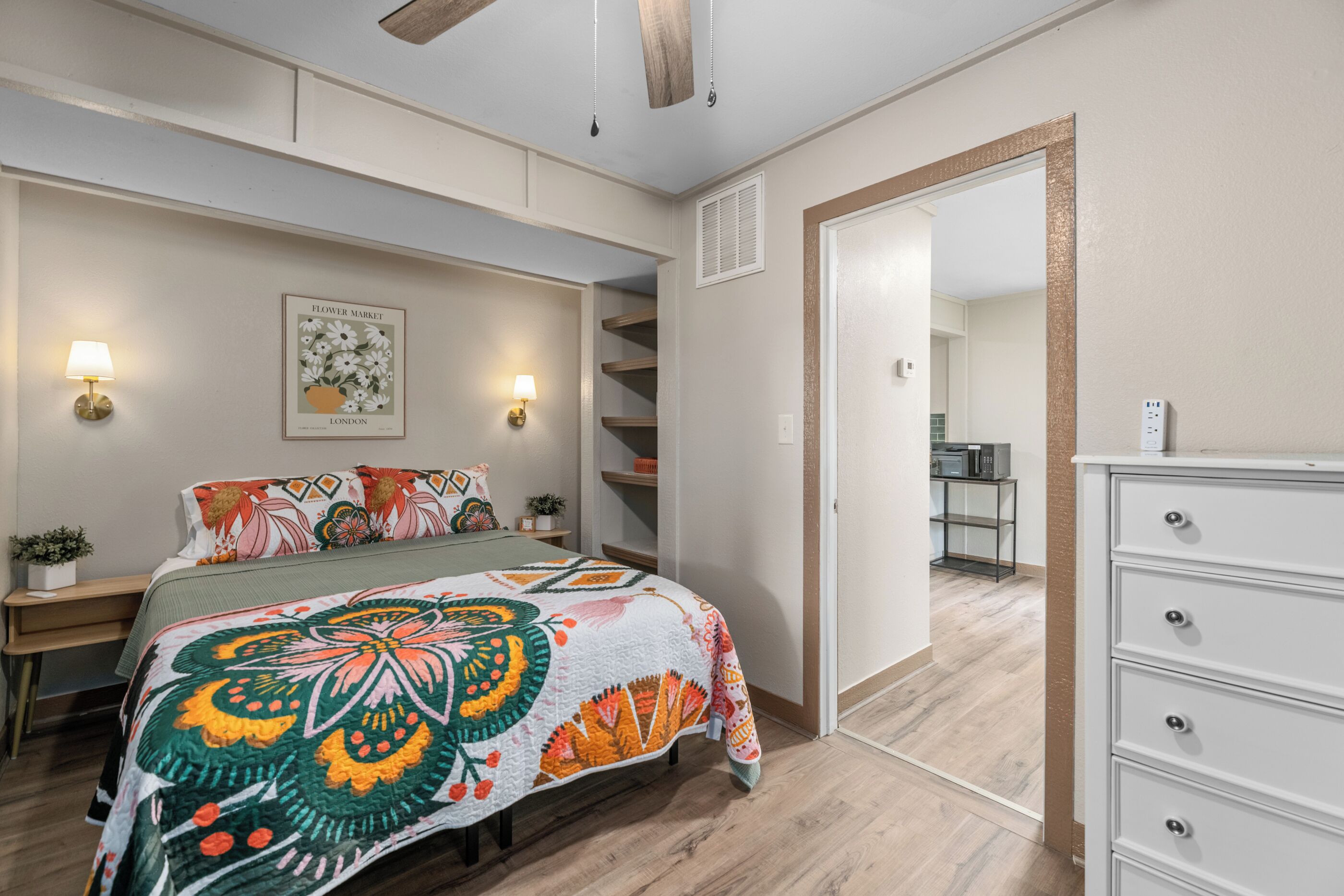 Une chambre à coucher comprenant un lit, des lampes de chevet, un tableau accroché au mur, une commode et une porte donnant sur la cuisine.