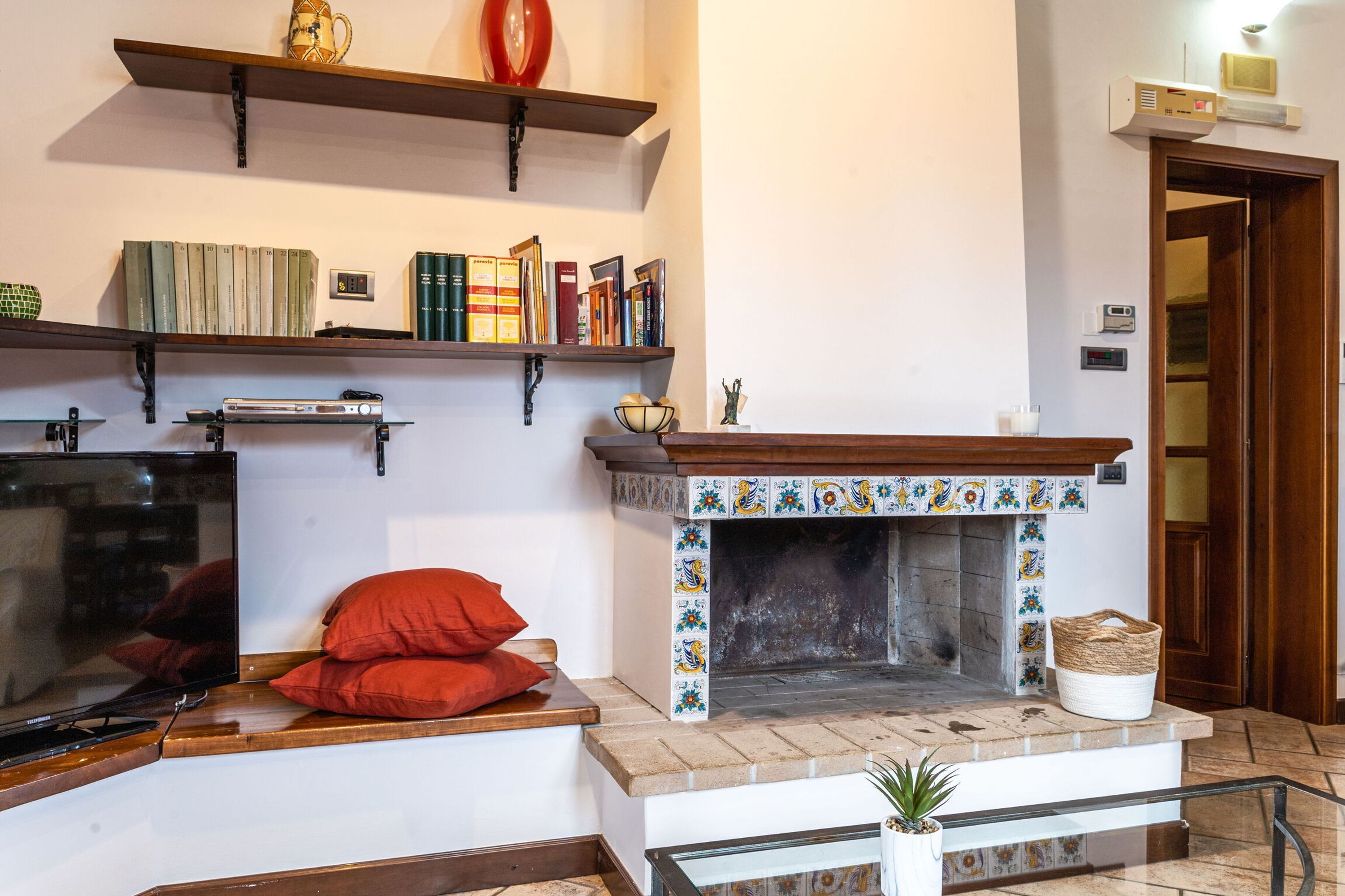 A living room with a fireplace, bookshelves, a television, and a red pillow on a bench.