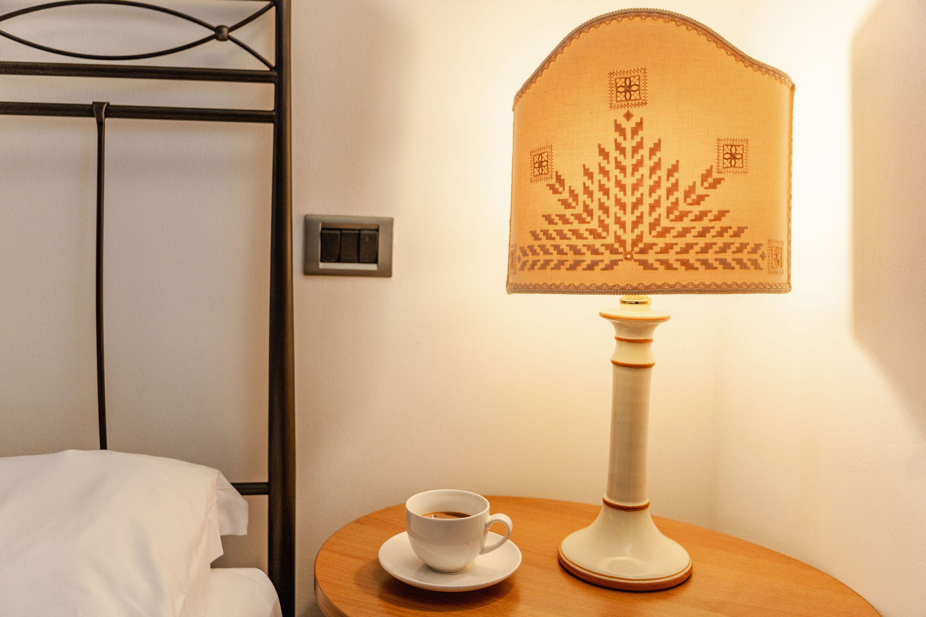 Ein Bett mit weißer Bettwäsche, ein Nachttisch mit einer Kaffeetasse und eine Lampe mit einem gemusterten Lampenschirm.