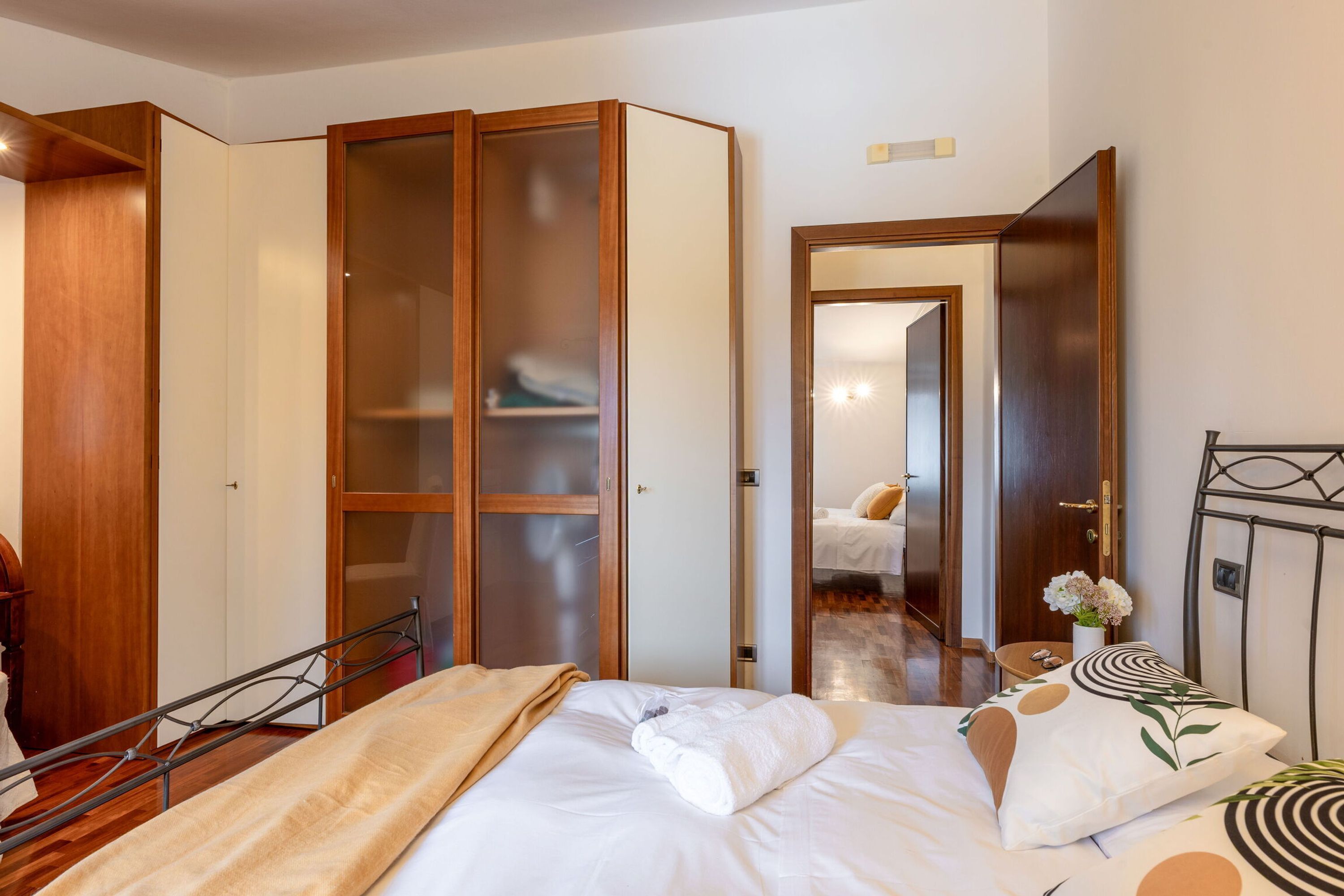 A hotel room with a bed, a wardrobe, a mirror, and a bedside table with a vase of flowers.