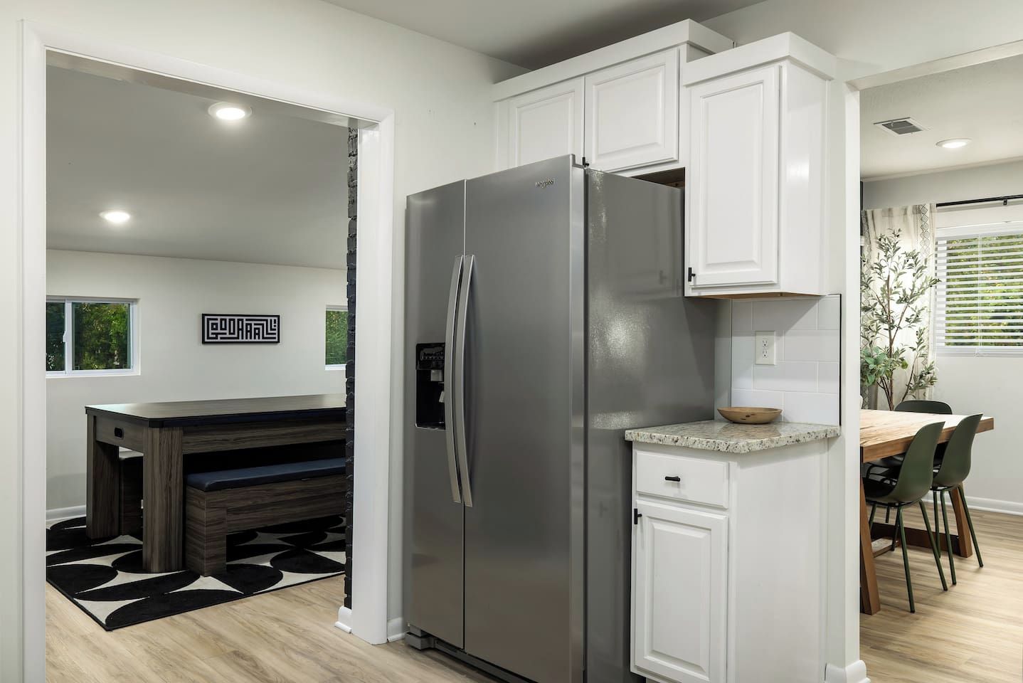 A modern kitchen with white cabinetry, a stainless steel refrigerator, a dining area with a wooden table, and a black and white rug.