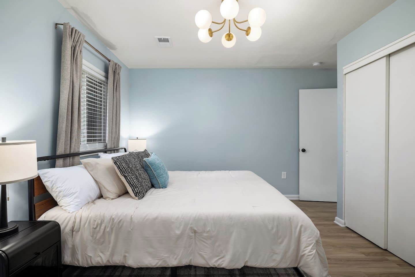 A bedroom with a bed, bedside lamps, a window with curtains, a door, and a chandelier.