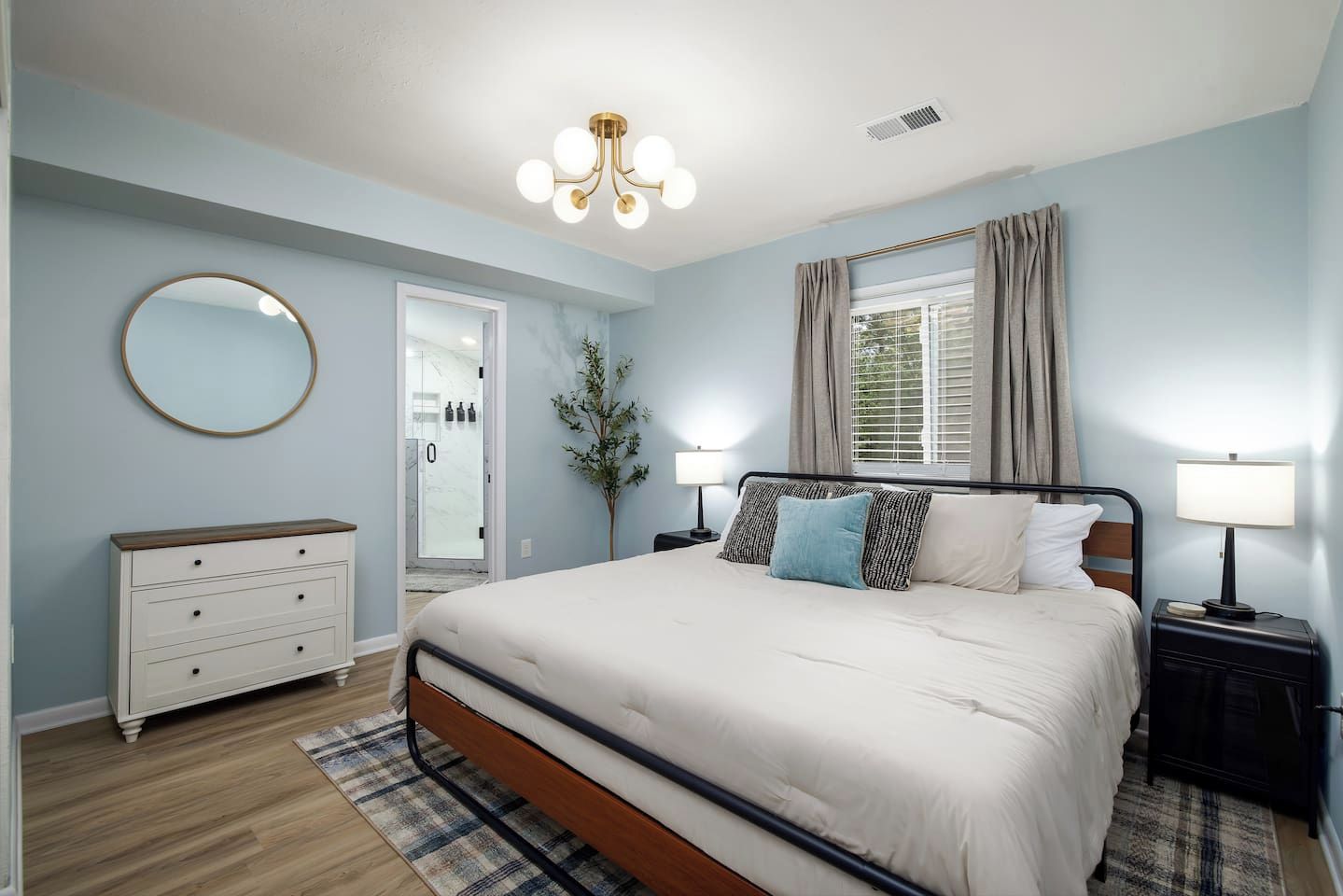 A bedroom with a bed, bedside tables, a dresser, a mirror, a chandelier, and a window with curtains.