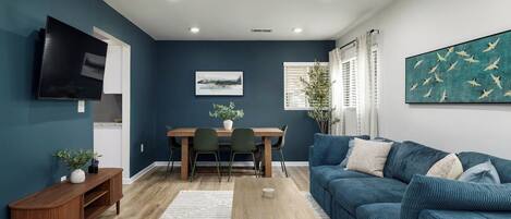 A modern living room with a dark blue wall, a wooden dining table, a blue sofa, and a flat-screen TV mounted on the wall.