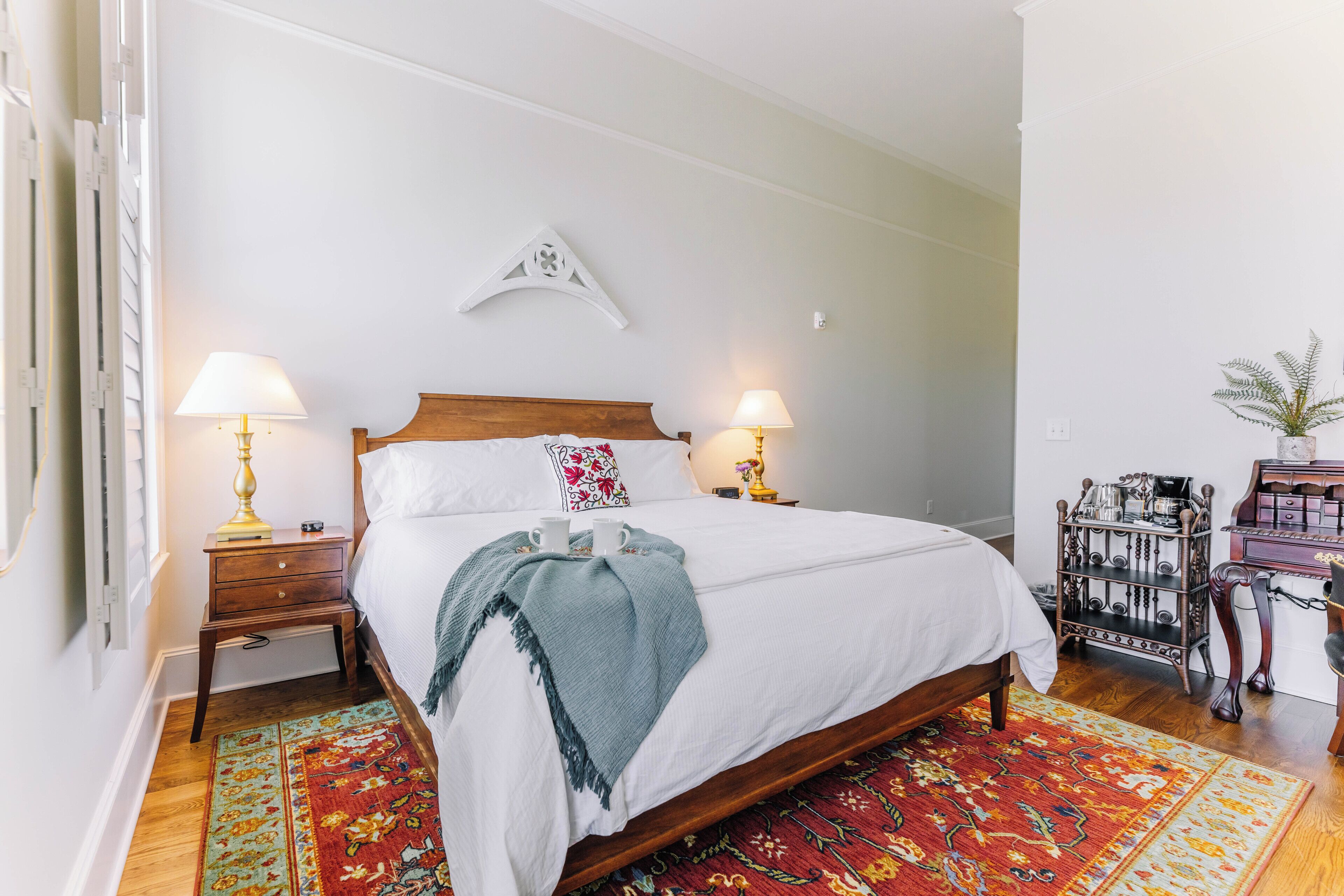 A neatly made bed with two bedside lamps, a decorative wall piece, and a coffee station with a plant.