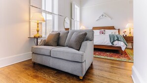 A modern bedroom with a grey sofa, a wooden floor, a bed with a white duvet, and a large window.