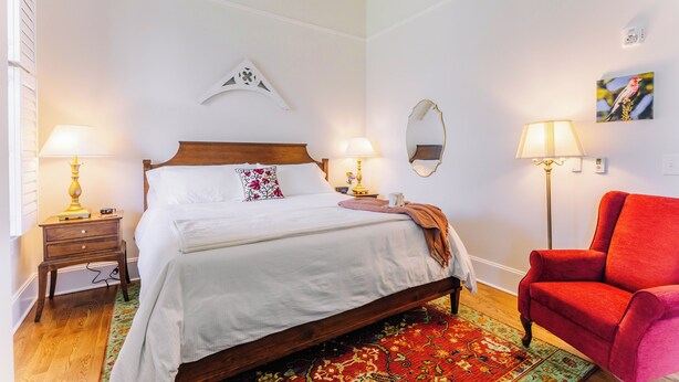 Une chambre à coucher bien rangée, avec un lit en bois, deux tables de chevet avec des lampes, un fauteuil rouge, un miroir et une œuvre d’art accrochée au mur.