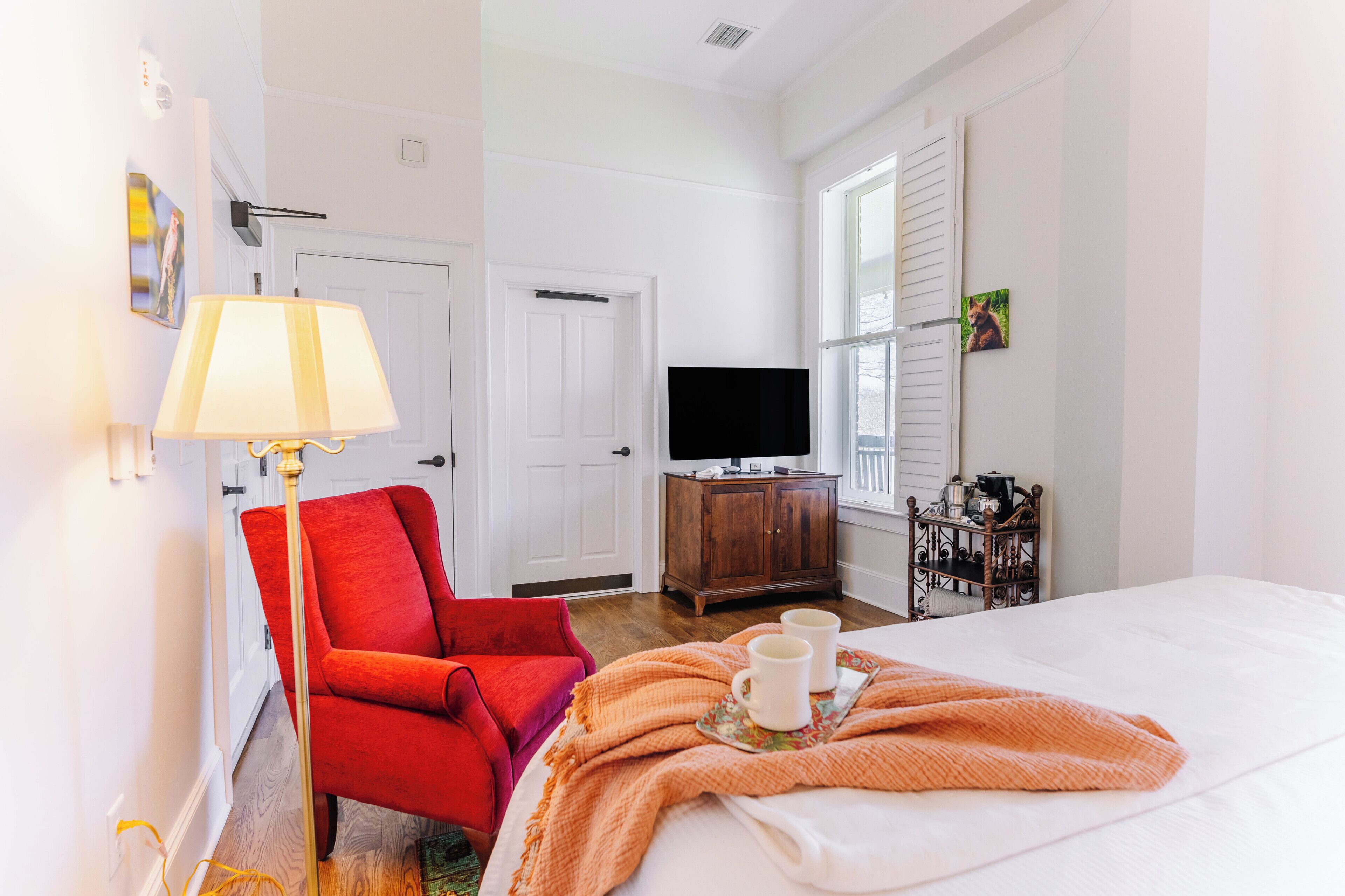 Une chambre d’hôtel avec un lit, un fauteuil rouge, une télévision, une armoire en bois et une fenêtre avec des volets.