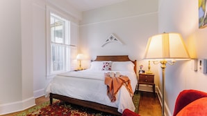 Une chambre à coucher avec un lit, des lampes de chevet, une table de chevet, un fauteuil rouge et une fenêtre avec des volets.